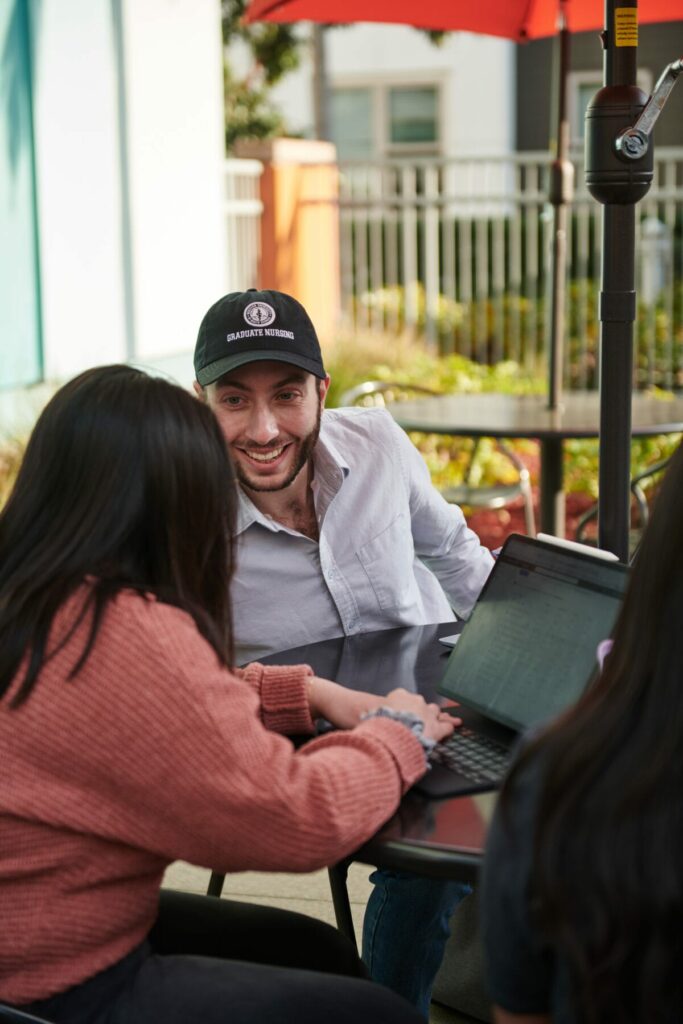 Three people sit around an outdoor table with laptops. One person, wearing a baseball cap and white shirt, is smiling and talking to another person in a pink sweater.