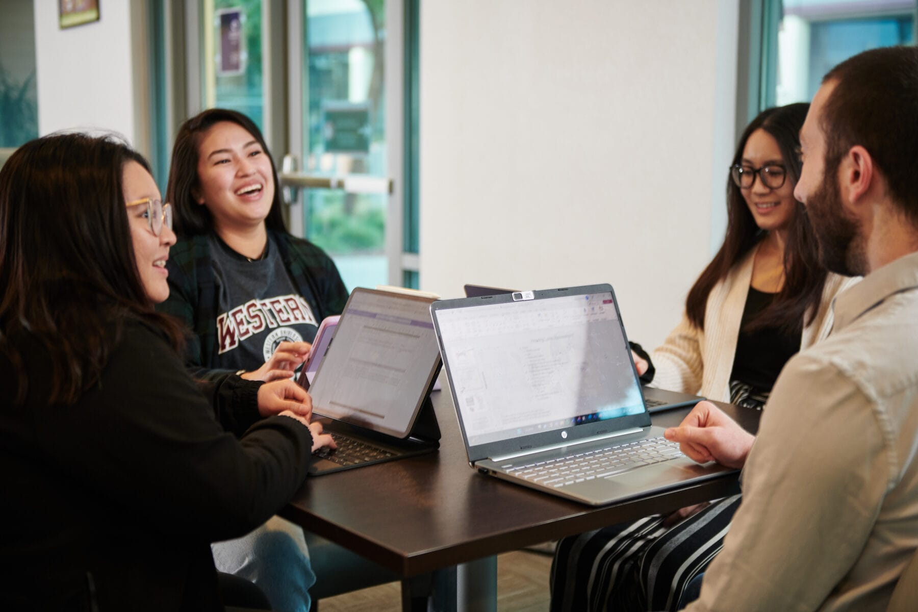 Four people sit around a table with laptops, engaged in conversation and smiling in a bright indoor setting.