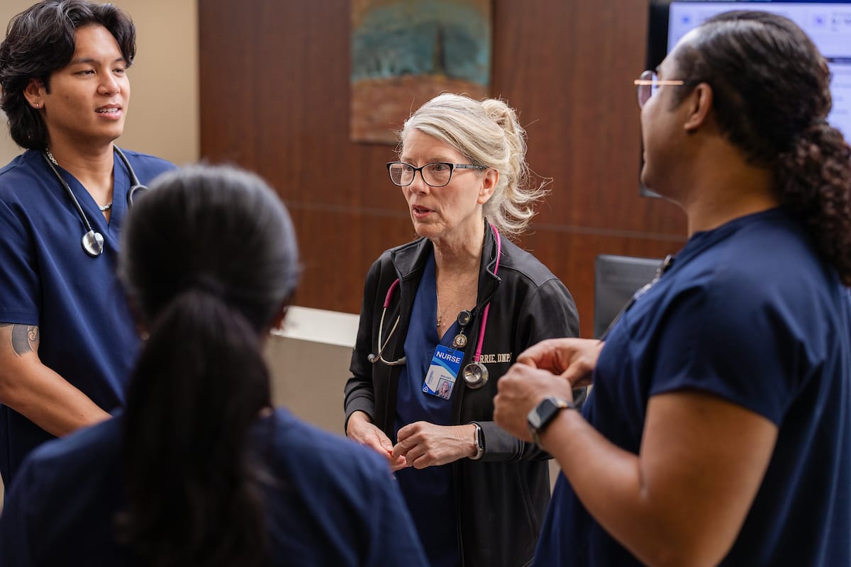 Four healthcare professionals in scrubs, including one with a stethoscope and name badge, stand in a hospital setting having a discussion.