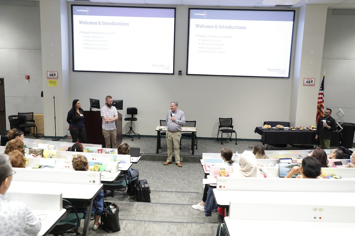 A man stands speaking at the front of a lecture hall, flanked by two people. The audience is seated, and slides on two screens read 