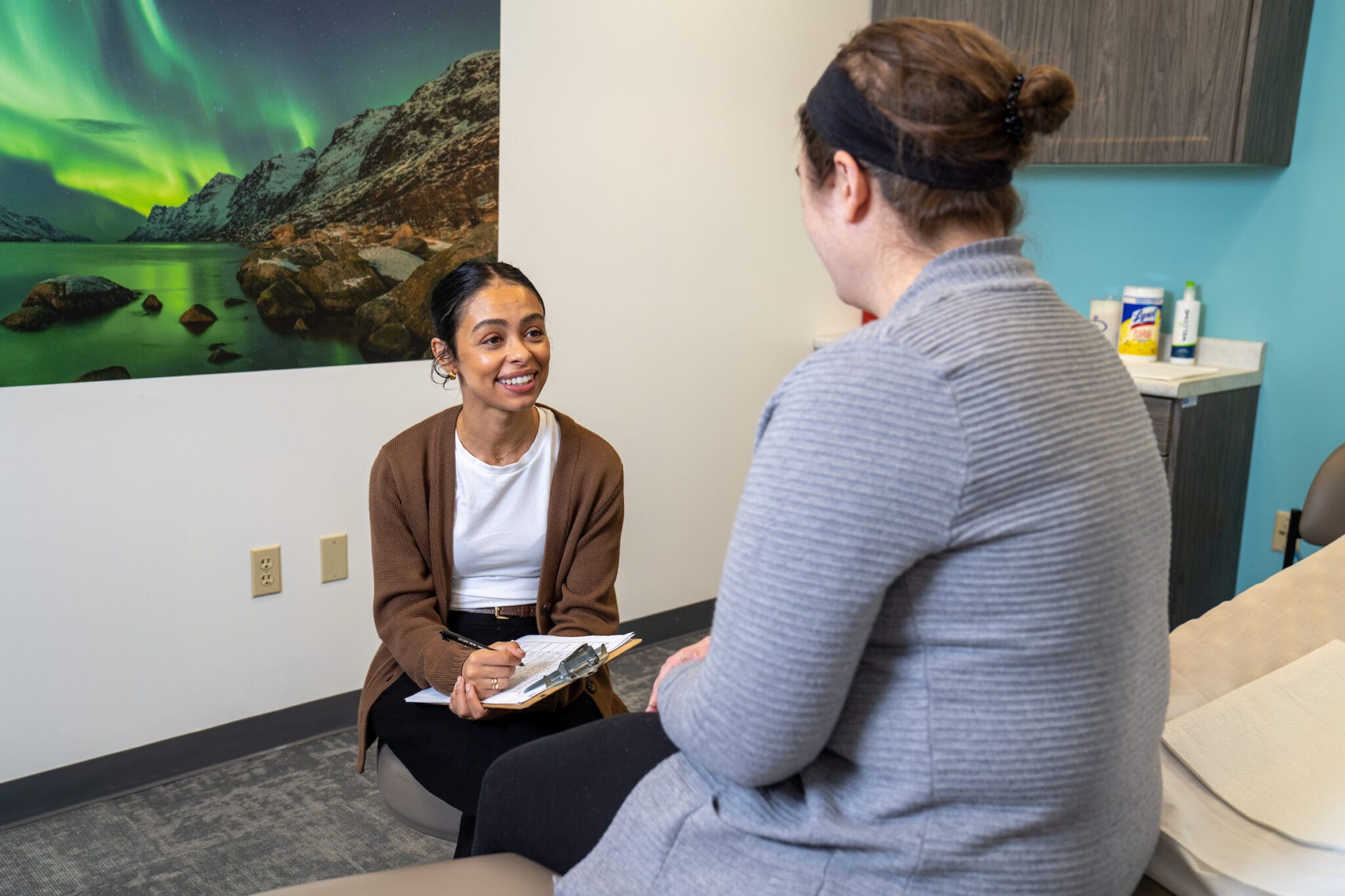A healthcare professional sits with a clipboard, smiling and speaking with a patient in a medical office with a nature photo on the wall.