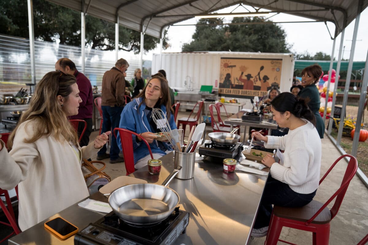 Two women interact near a table with cooking supplies in an outdoor setting; a large sign behind them reads, “We can feed ourselves together.”.