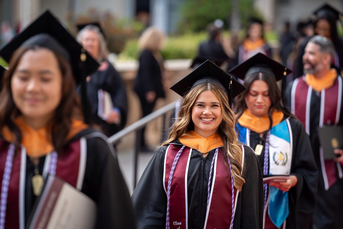Group of graduates in caps and gowns walking in a procession, focusing on a smiling woman in the center.