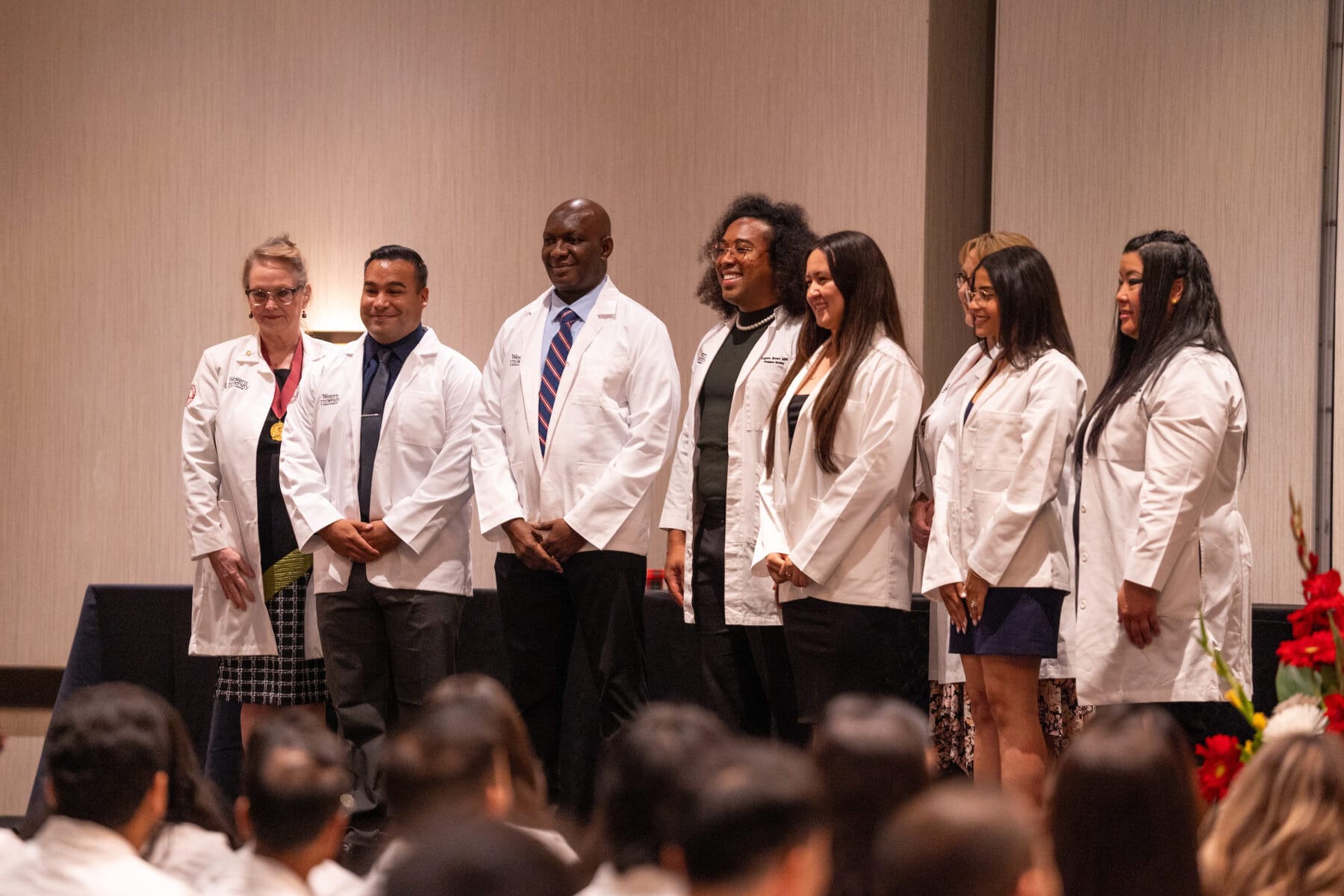Eight individuals in white coats pose for a photo in a formal setting. Six women and two men stand in a line, facing forward, with some smiling. Red floral arrangements are visible in the background.