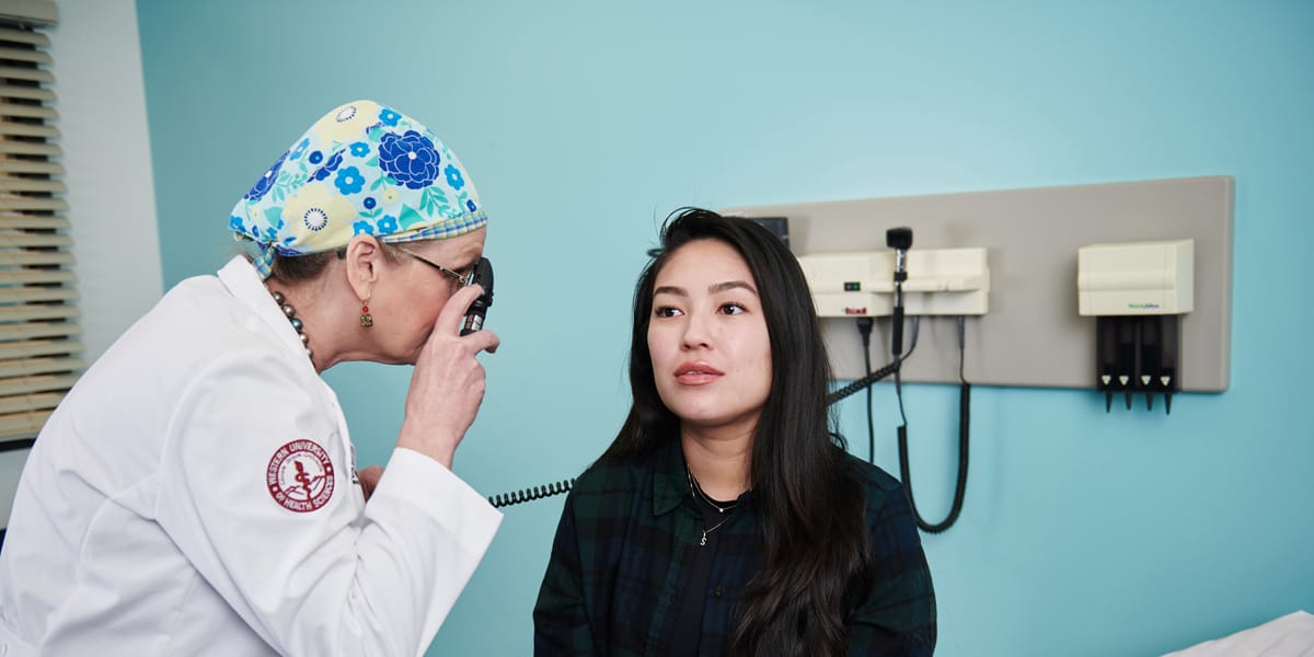 A doctor examines a patient's eye with an ophthalmoscope in a medical exam room. The patient sits on the exam table facing forward.