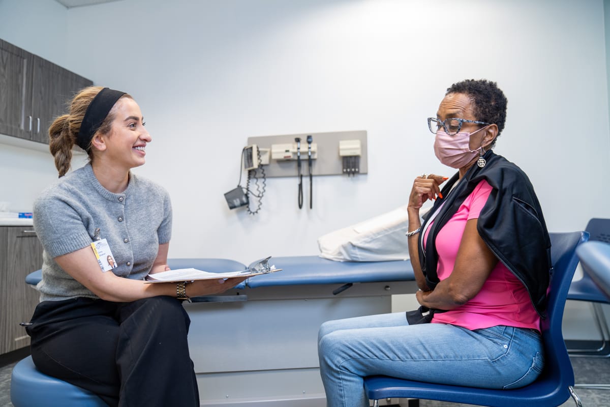 A healthcare worker and a seated woman wearing a mask converse in a medical examining room.