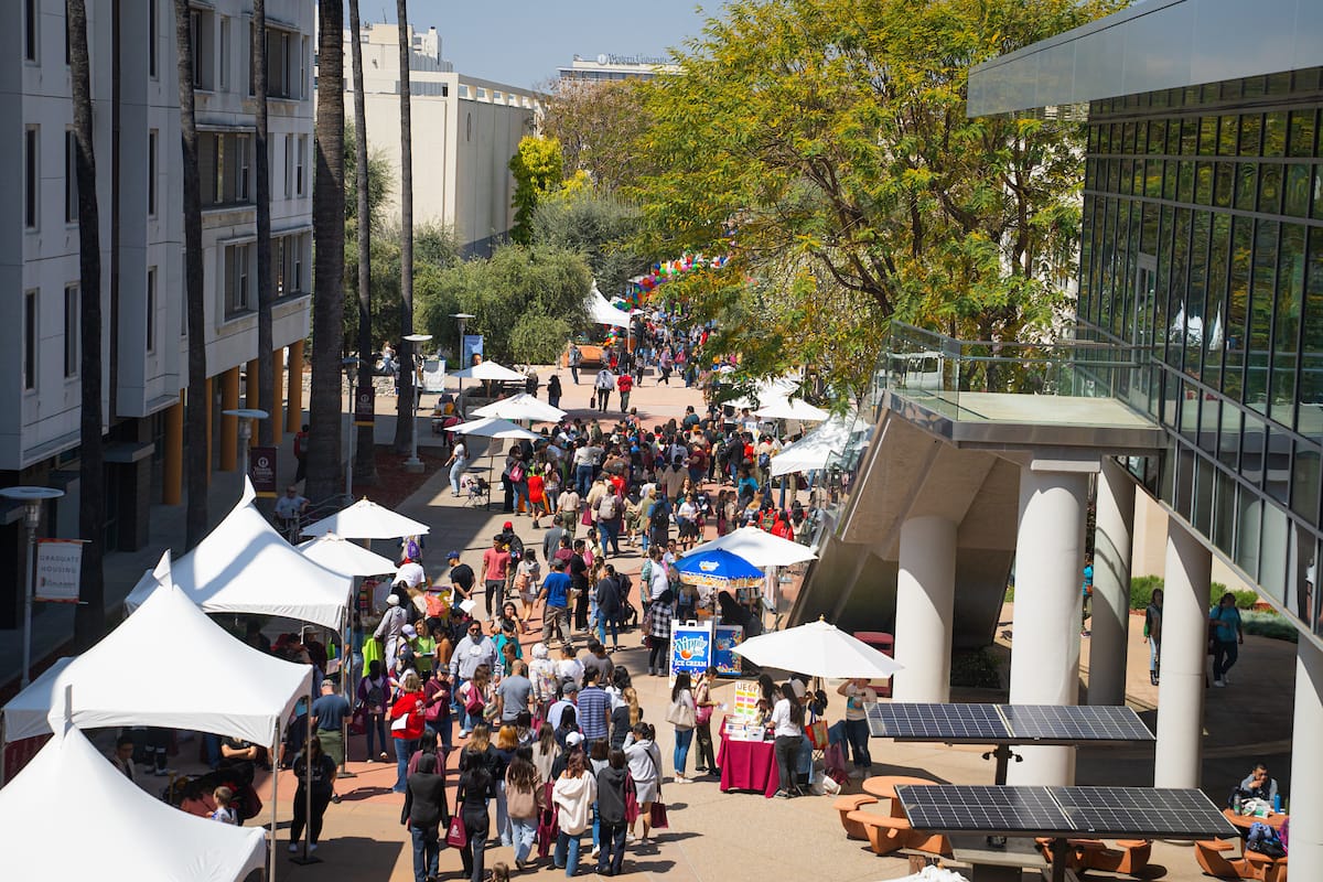A crowded outdoor event with people walking between white canopy tents in a plaza area, adjacent to modern buildings and a glass-walled structure. Trees and greenery are present. Clear, sunny day.