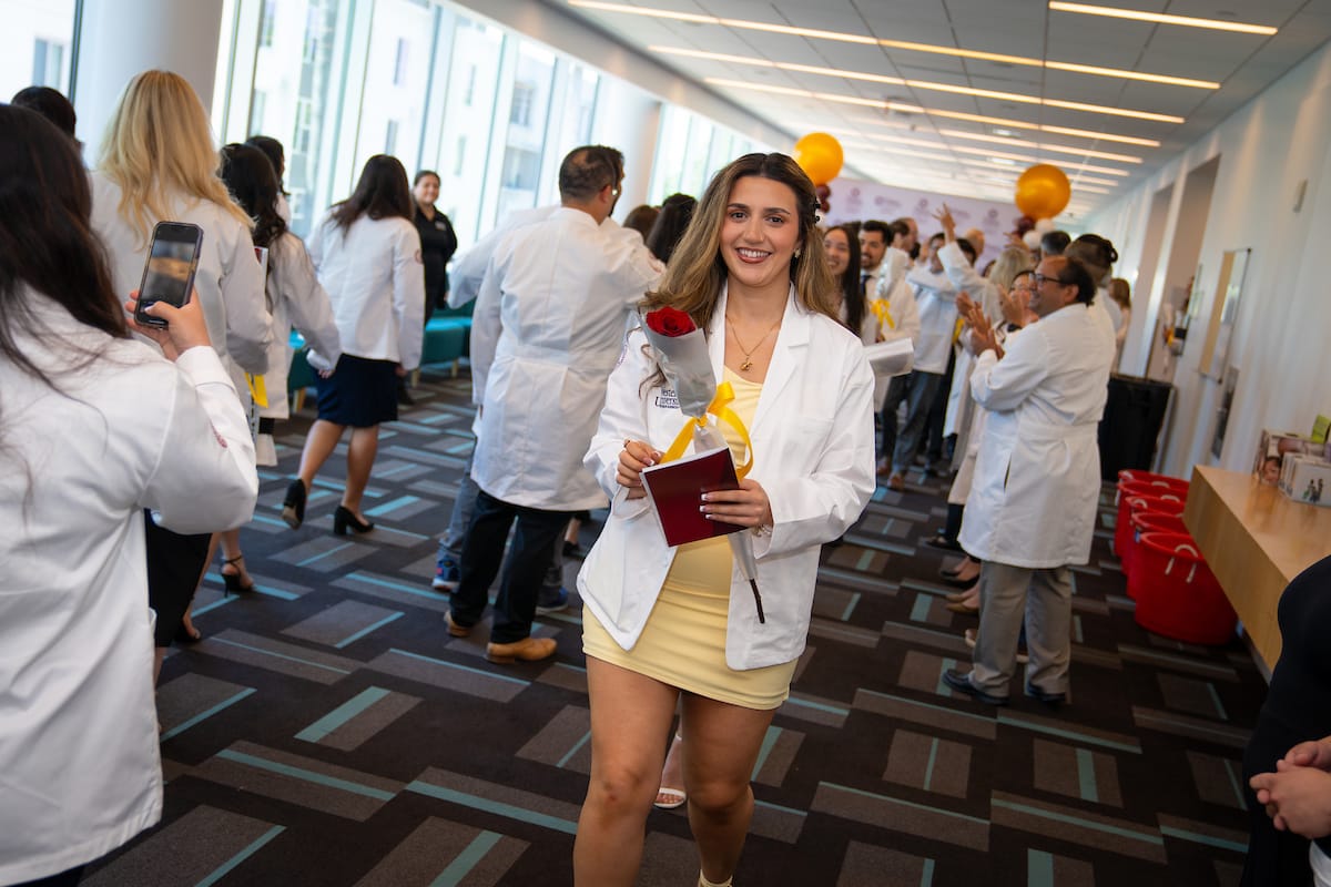 A person wearing a white coat holds a red rose and a booklet while smiling and walking down a corridor, surrounded by others in white coats.