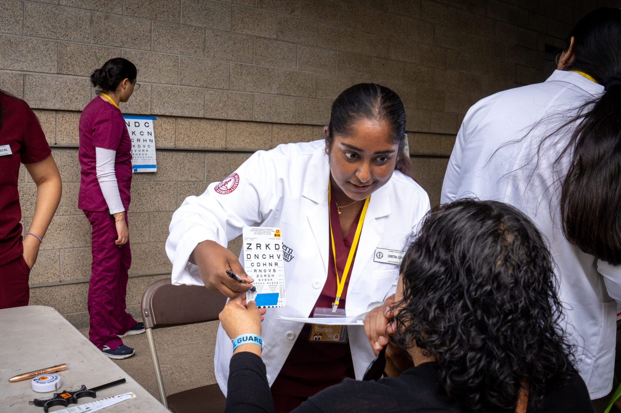 A healthcare worker in a white coat conducts an eye test for a seated patient, while others assist nearby in a clinical setting.