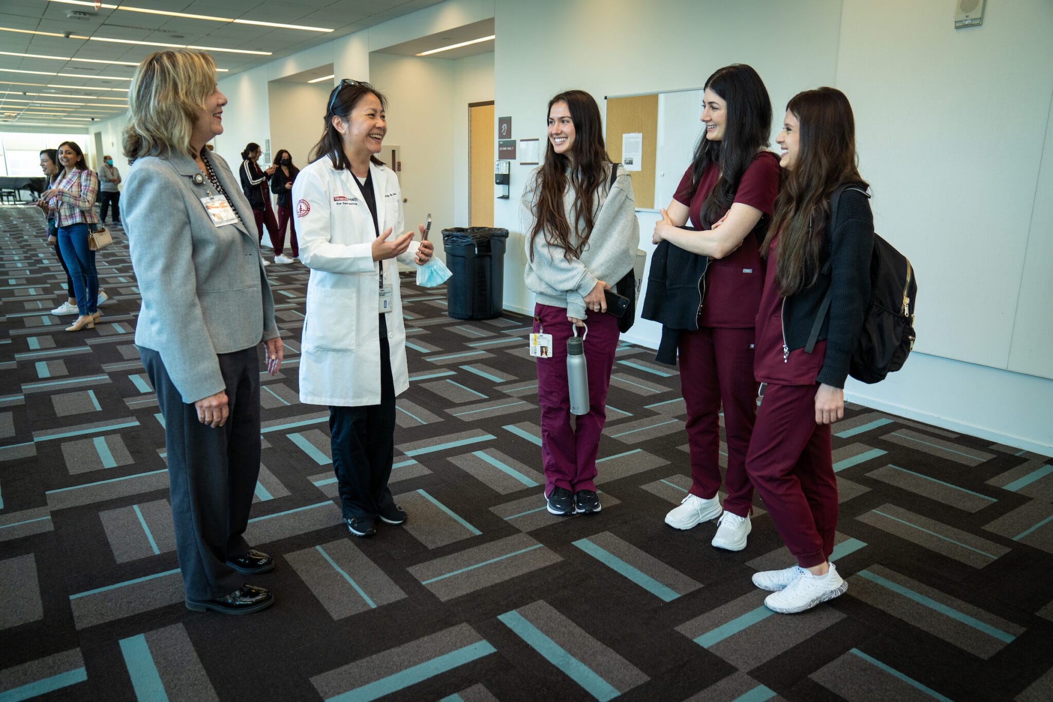 A group of three students in maroon scrubs and two women in professional attire stand talking in a hallway with patterned carpet.