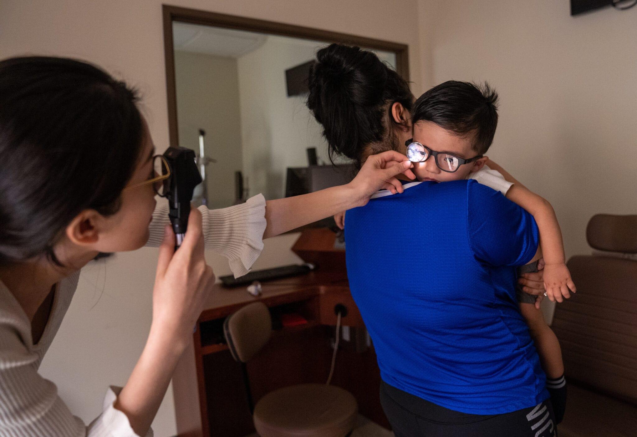 A healthcare professional examines a young boy’s eyes with an ophthalmoscope while he is held by an adult in a clinic room.