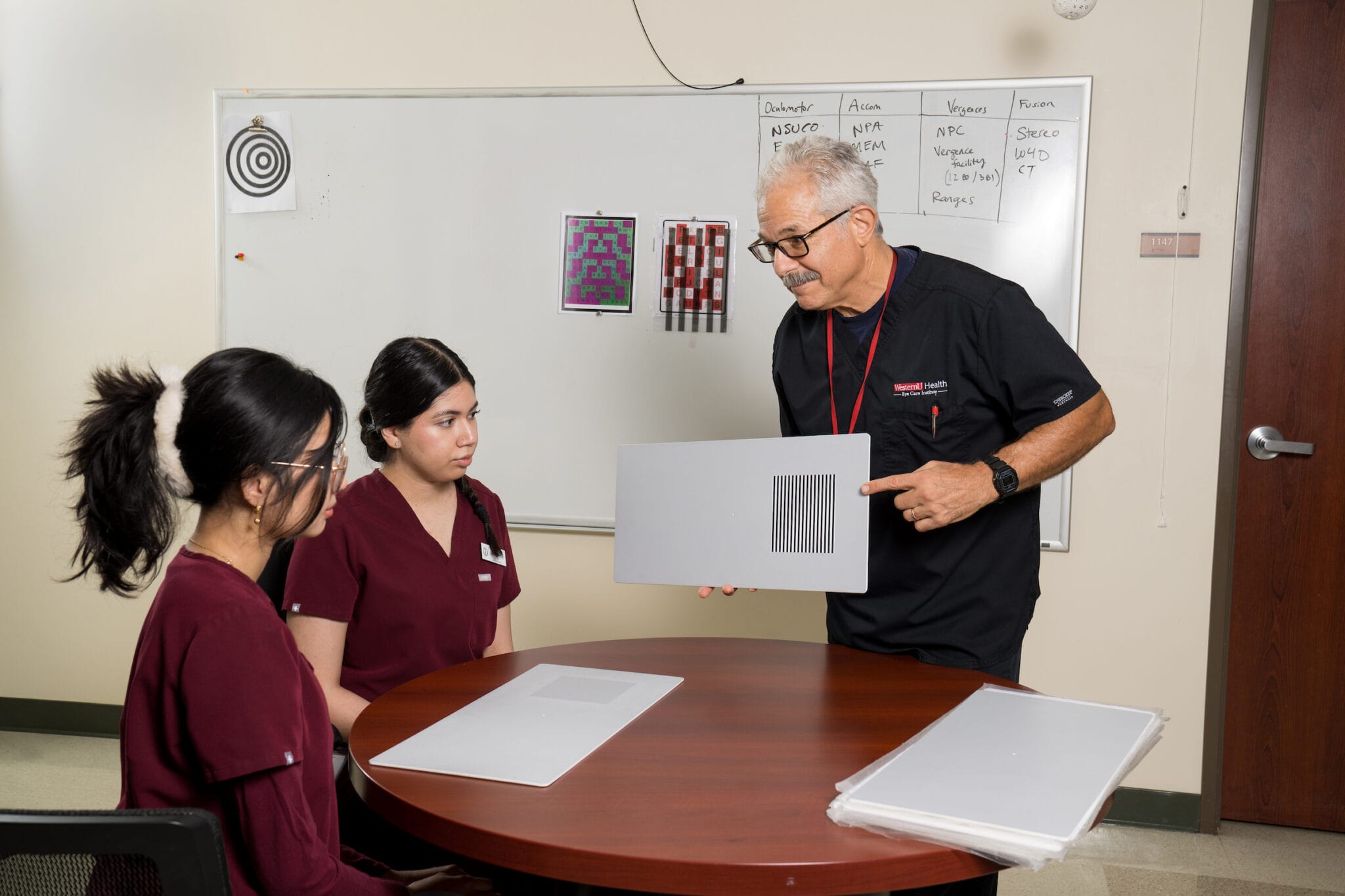 An instructor shows two students a test pattern sheet in a classroom with a whiteboard and target charts in the background.