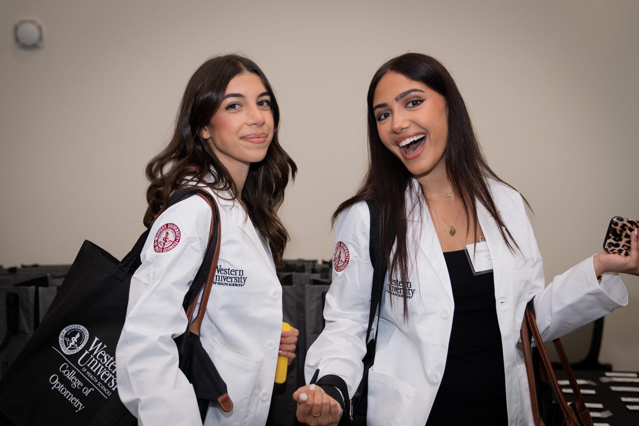 Two women wearing white lab coats with Western University logos, smiling and holding bags, stand indoors in front of a neutral background.