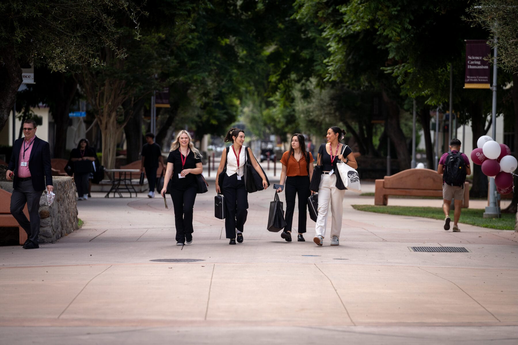 A group of people walking on a tree-lined pathway, some carrying bags, with benches and balloons in the background.