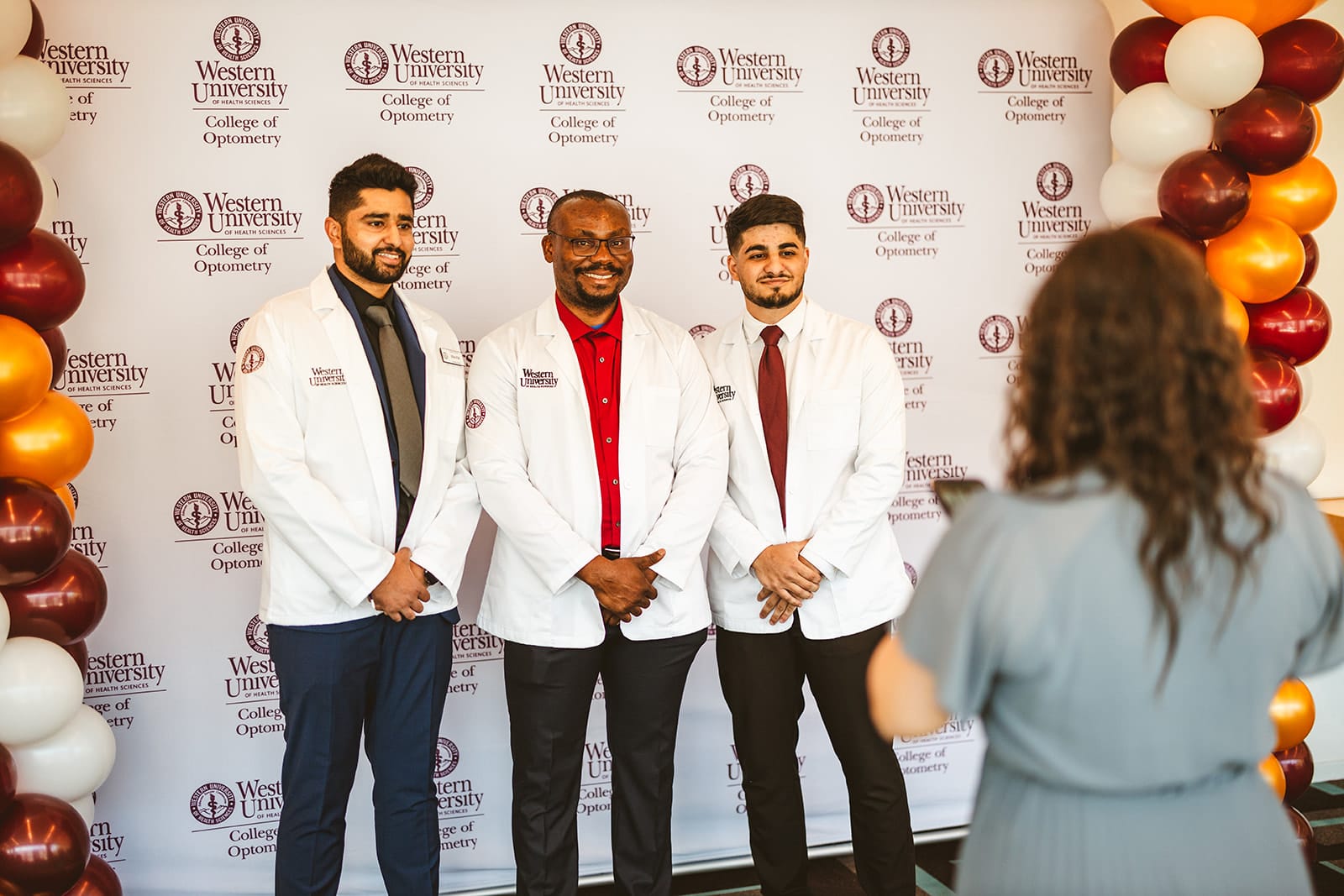 Three individuals in white coats pose for a photo in front of a Western University College of Optometry backdrop, with balloons on either side and a person taking their picture.