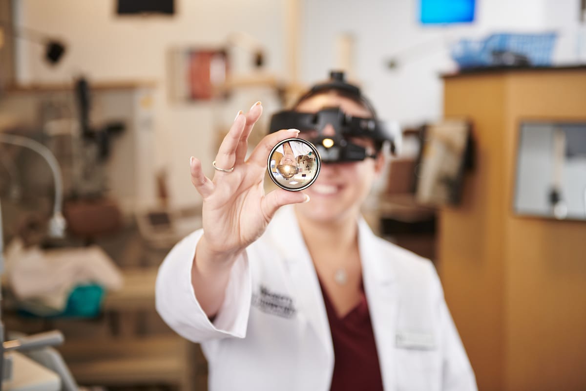 A person in a lab coat holds a lens while wearing a head-mounted magnifier in an optometrist's office.
