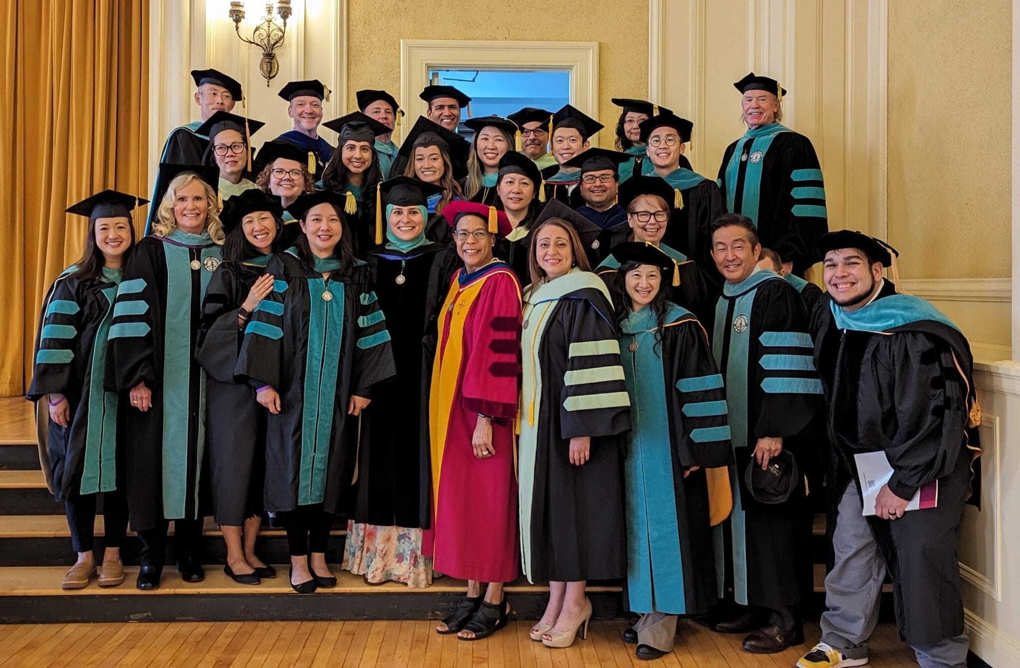 A group of people in academic regalia pose together indoors, smiling for a group photo at a graduation ceremony.