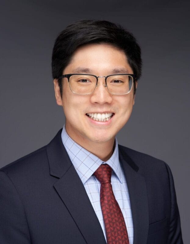 A man wearing glasses, a dark suit, a blue checkered shirt, and a red tie smiles at the camera against a plain dark background.