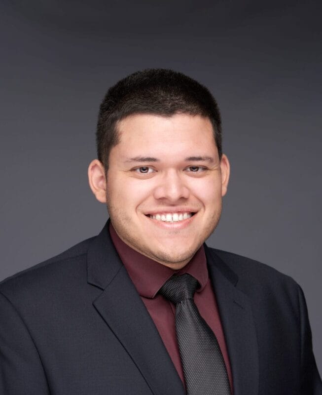 A man in a dark suit, maroon shirt, and black tie smiles at the camera against a plain gray background.
