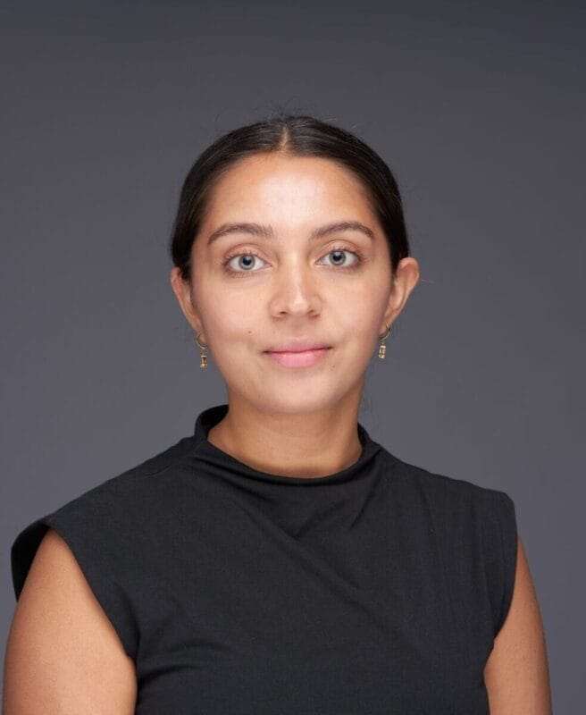 A woman with dark hair pulled back, wearing a sleeveless black top, stands in front of a plain grey background and looks directly at the camera.