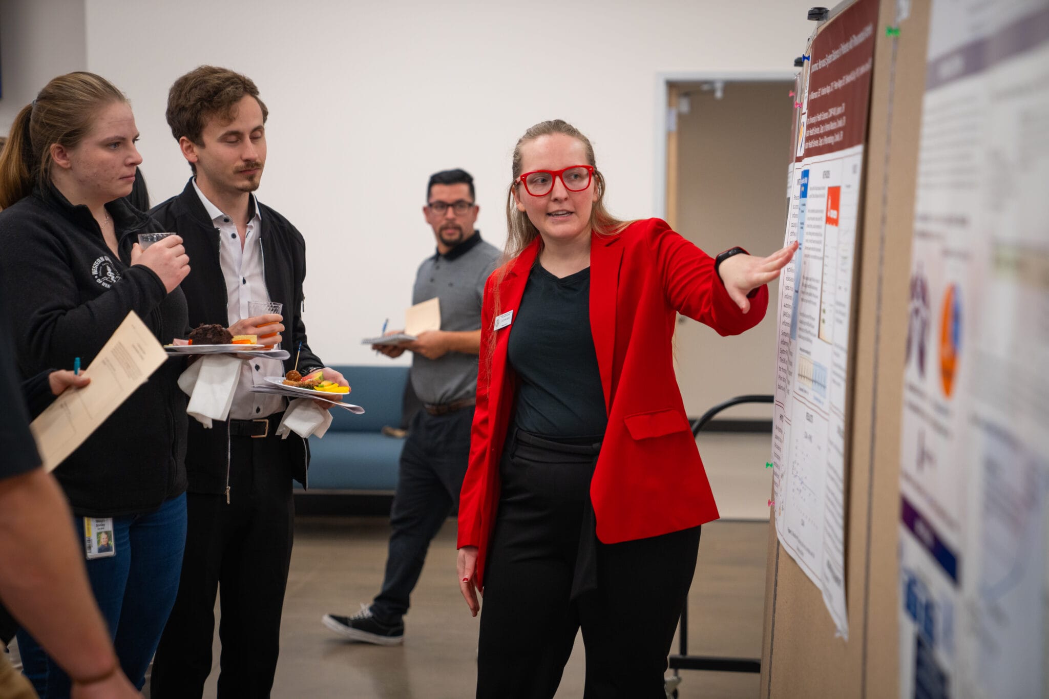 A woman in a red blazer presents information on a poster board to three people, two of whom are holding plates of food, in an indoor setting.