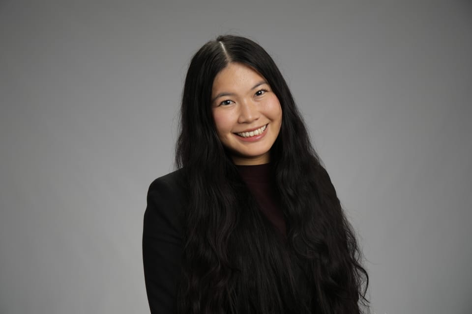 A woman with long black hair, wearing a black blazer and dark top, smiles in front of a plain gray background.