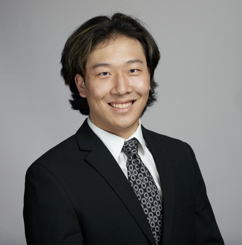 Young man wearing a black suit, white shirt, and patterned tie, smiling at the camera against a plain gray background.