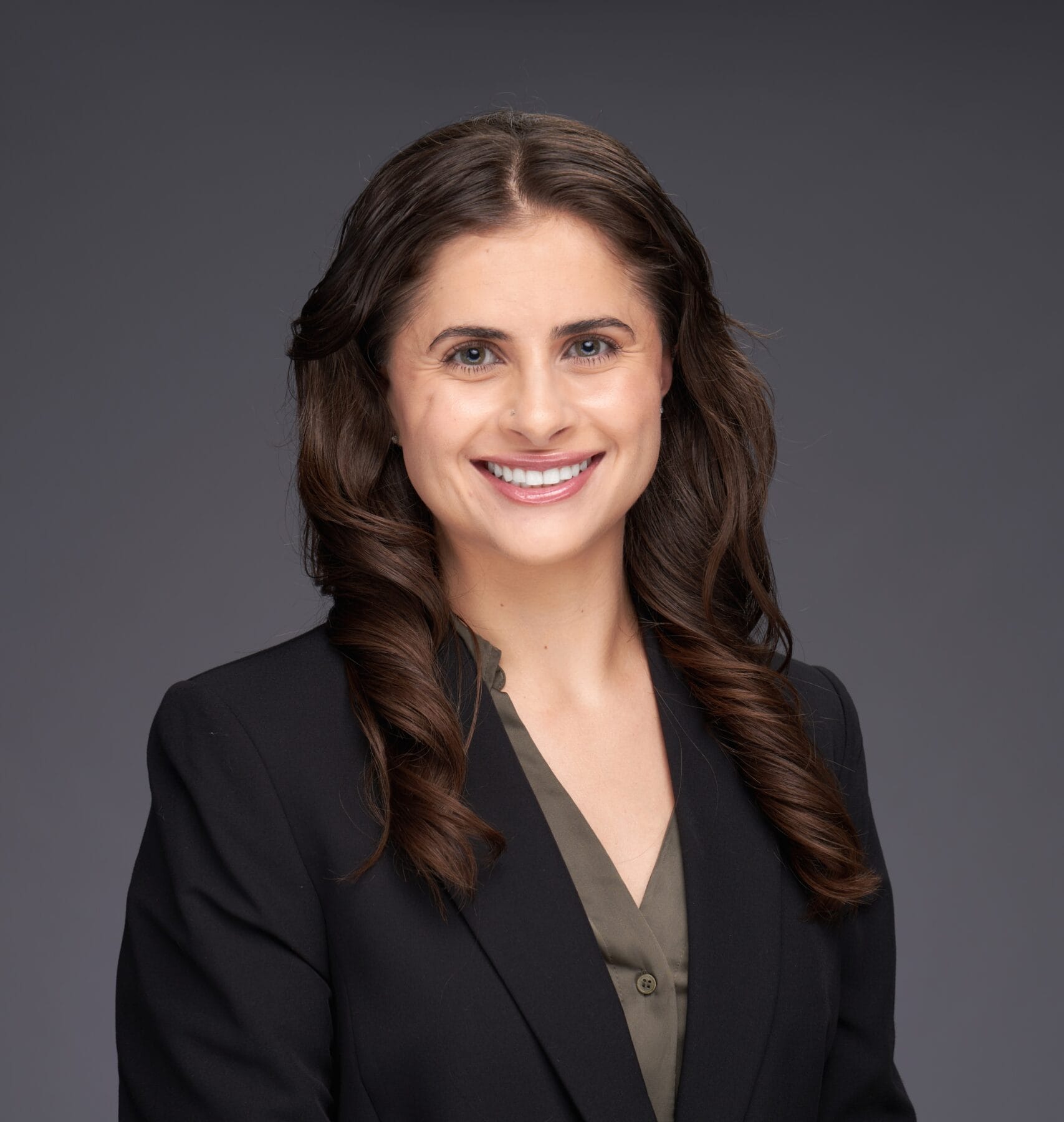 A woman with long brown hair wearing a dark blazer and olive blouse smiles at the camera against a plain gray background.