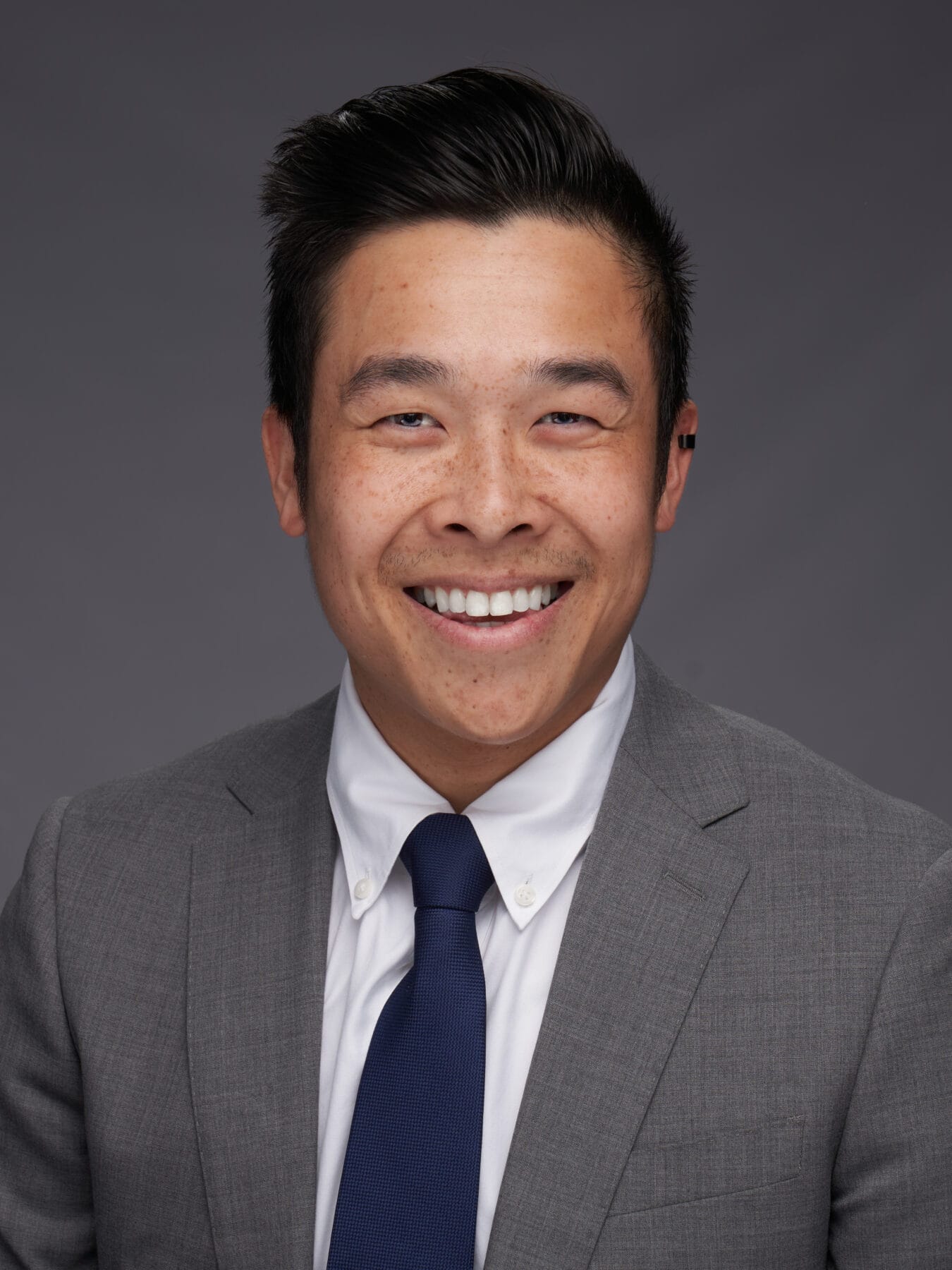 A man in a grey suit, white shirt, and navy tie smiles at the camera against a plain grey background.