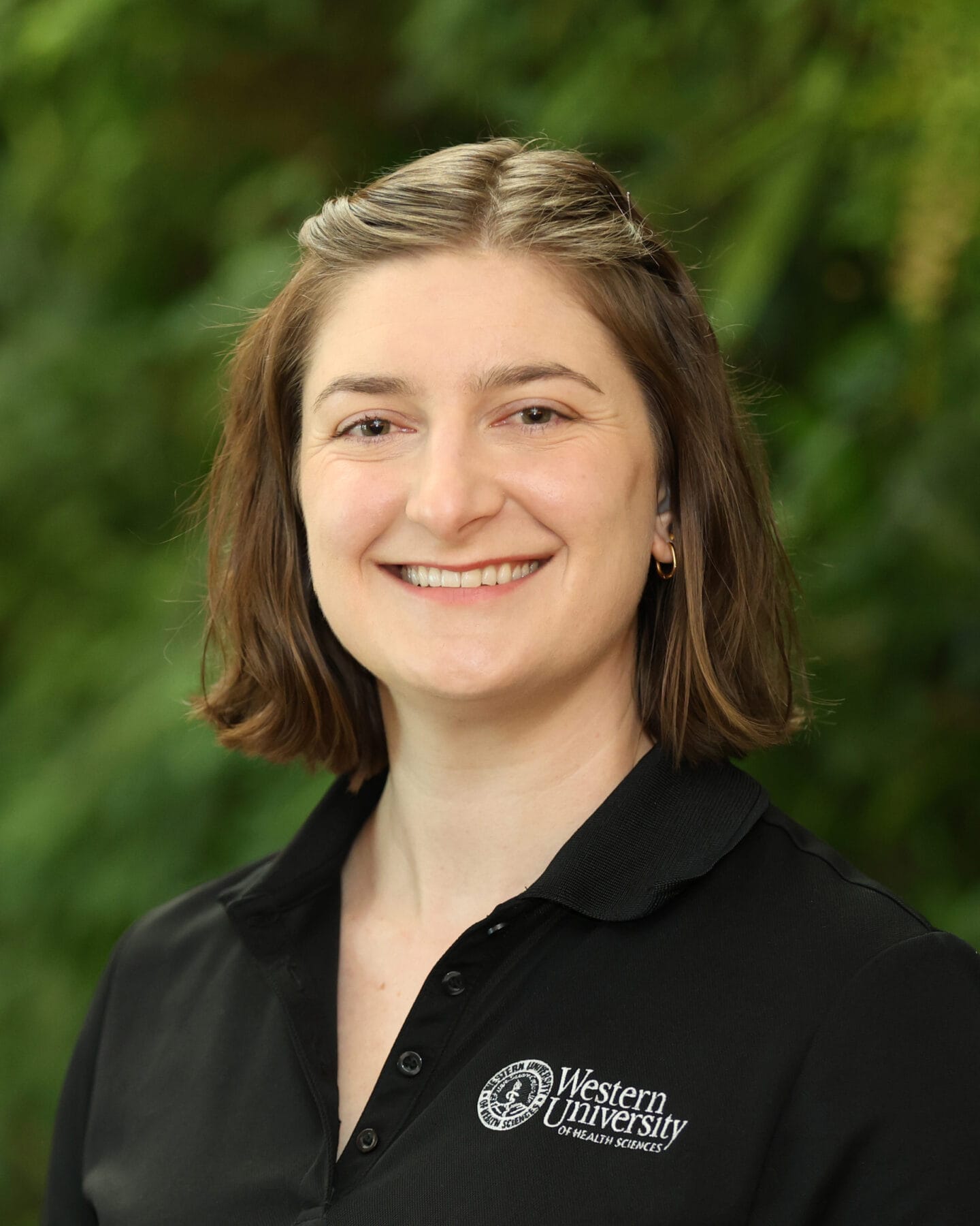 A woman with short brown hair wearing a black "Western University of Health Sciences" polo shirt smiles in front of a green, blurred background.