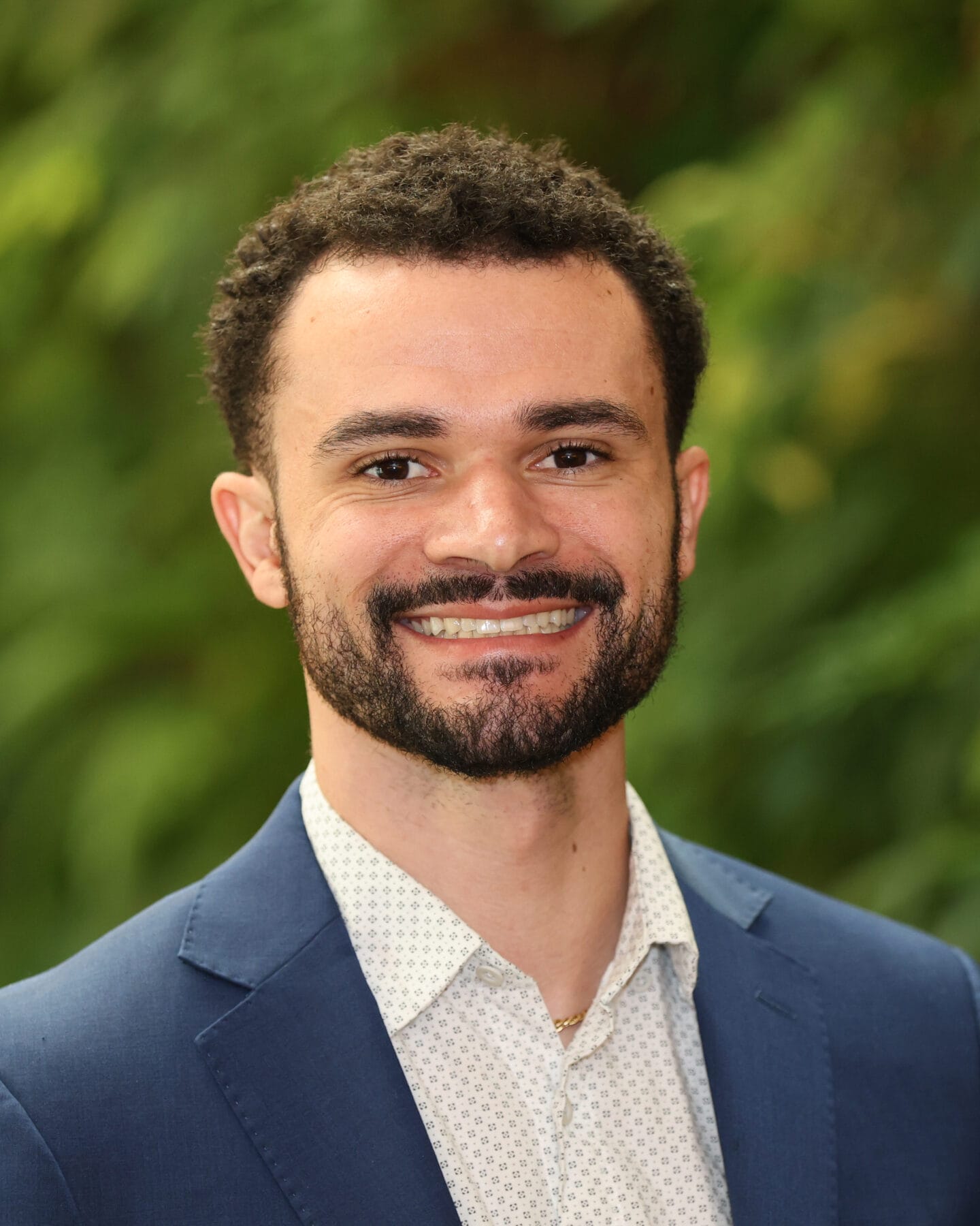 A man with short curly hair and a beard, wearing a blue suit jacket and white shirt, smiles in front of a blurred green background.