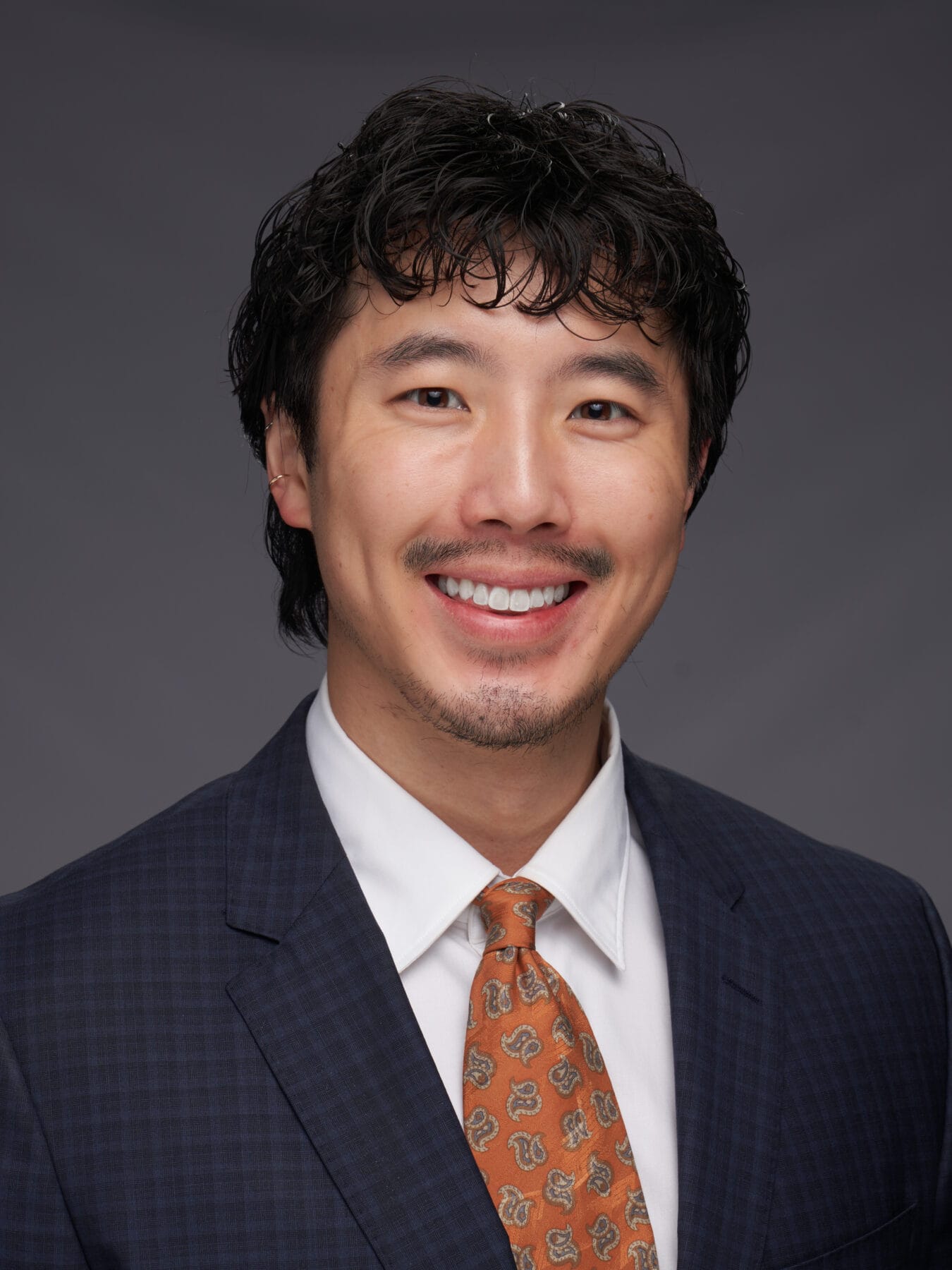 A man in a blue checkered suit, white shirt, and patterned orange tie smiles at the camera against a plain gray background.