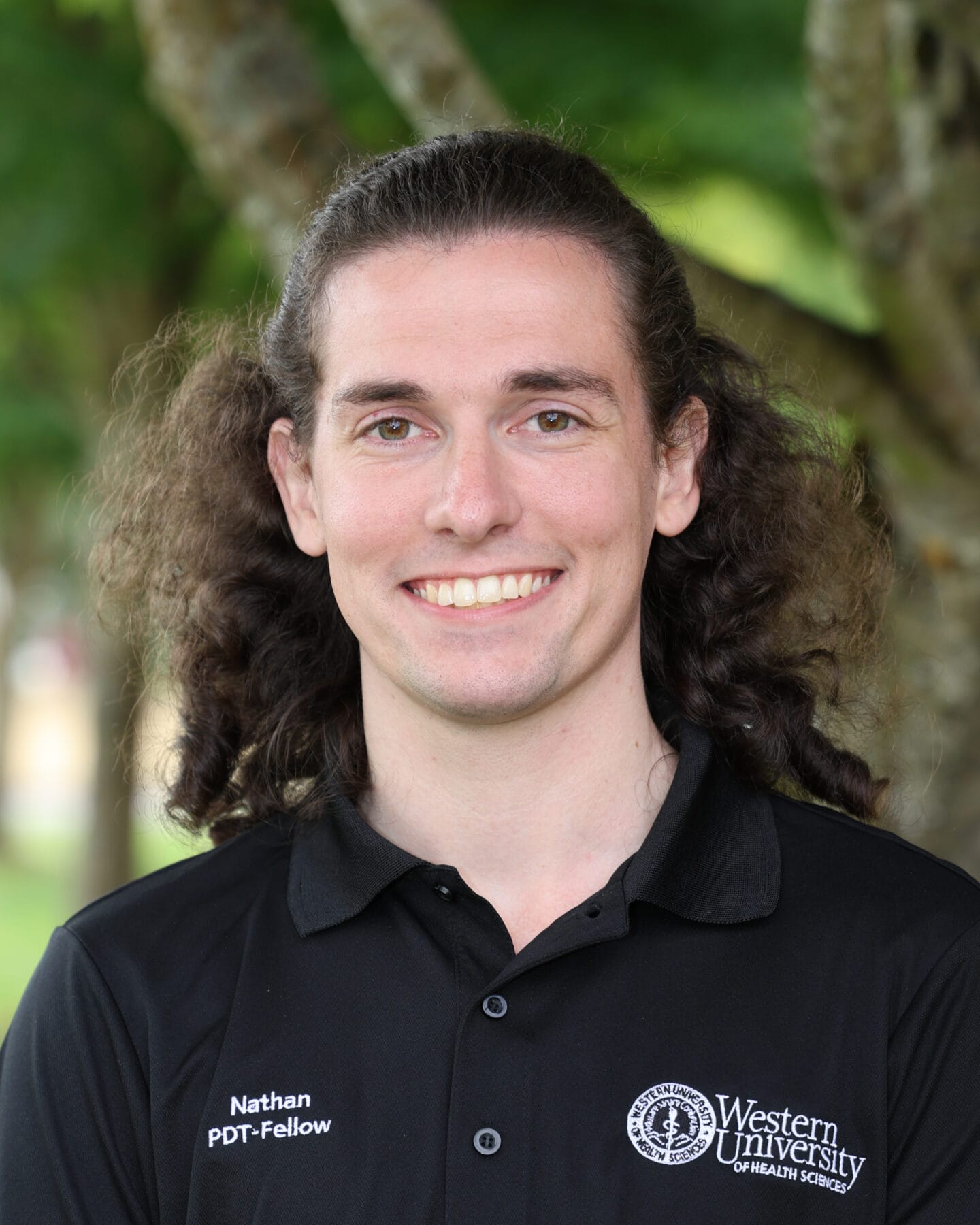 A person with long brown hair smiles at the camera, wearing a black "Western University of Health Sciences" polo shirt with the name "Nathan" embroidered on it. Trees are blurred in the background.