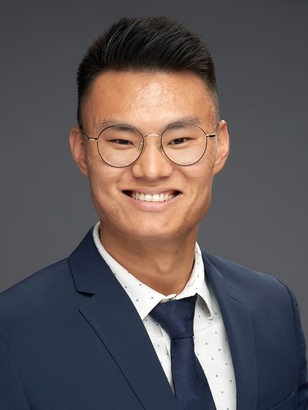 A man wearing glasses and a navy suit with a white shirt and tie smiles against a gray background.
