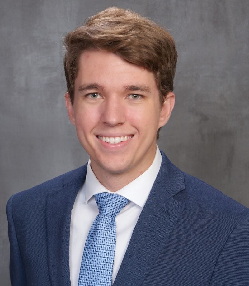 A young man with short brown hair wearing a blue suit, white shirt, and blue patterned tie, smiling in front of a gray background.
