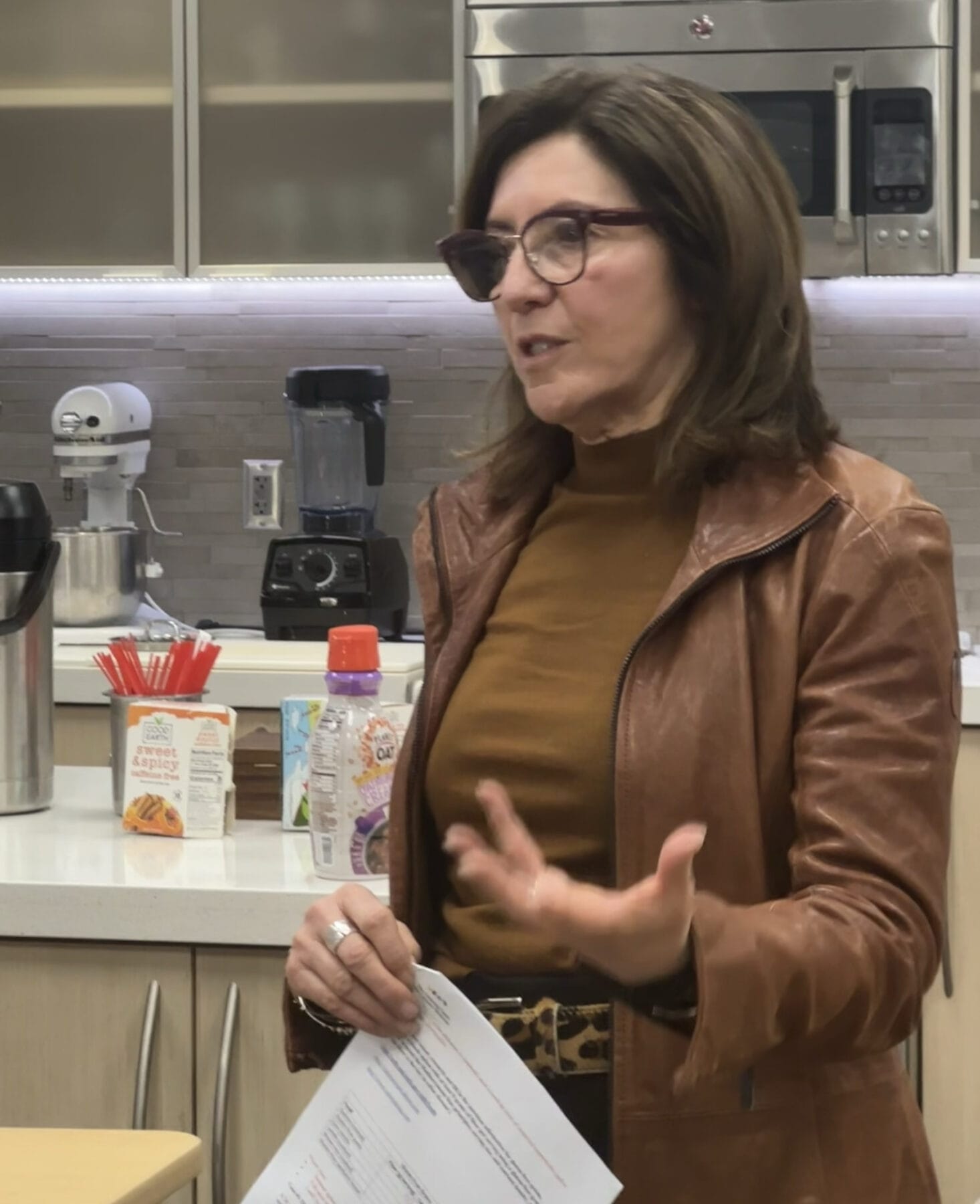 A woman in glasses and a brown jacket speaks while holding papers in a kitchen setting with appliances and coffee supplies in the background.