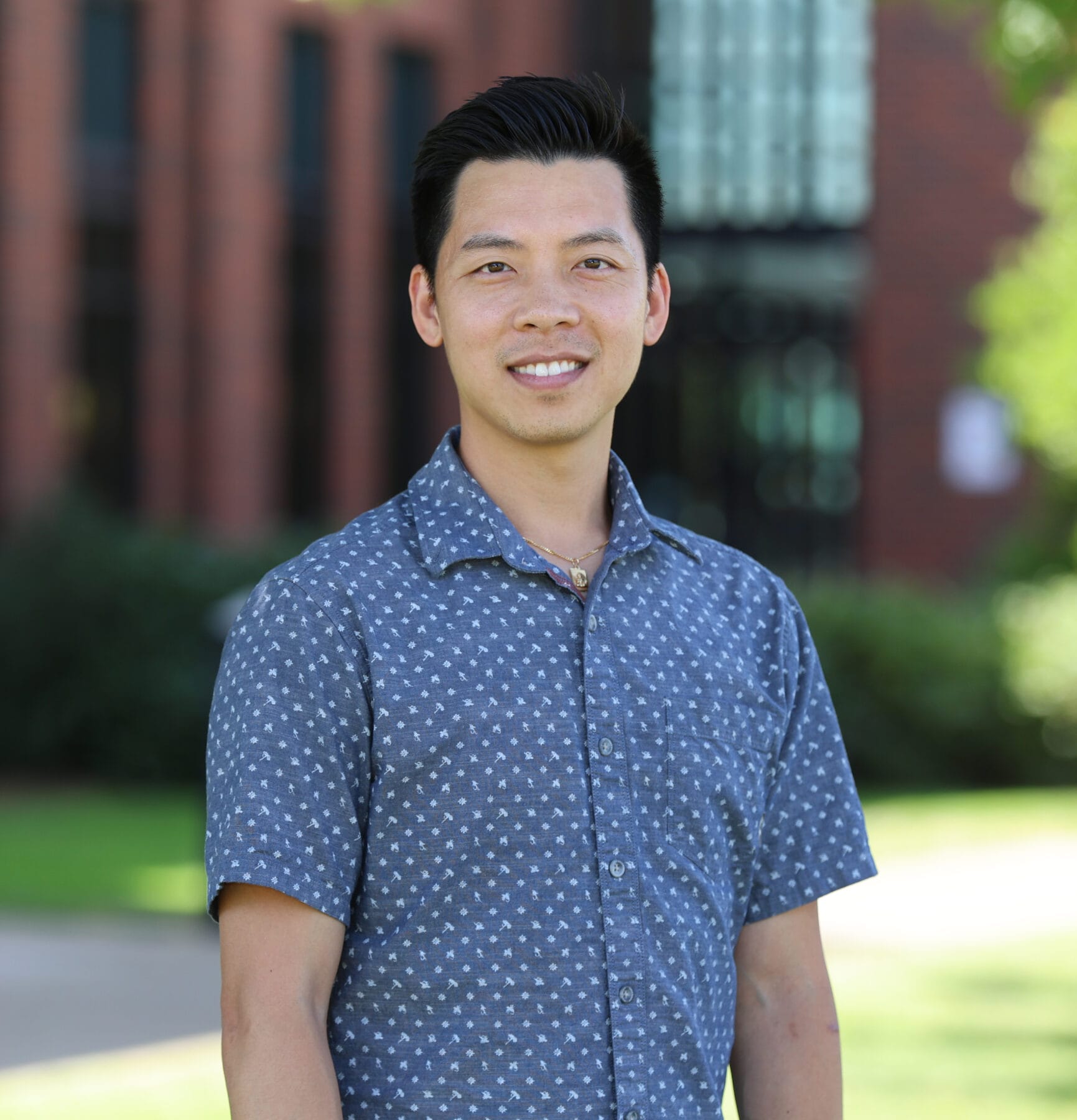 A person wearing a blue patterned shirt smiles while standing outdoors in front of a brick building and greenery.