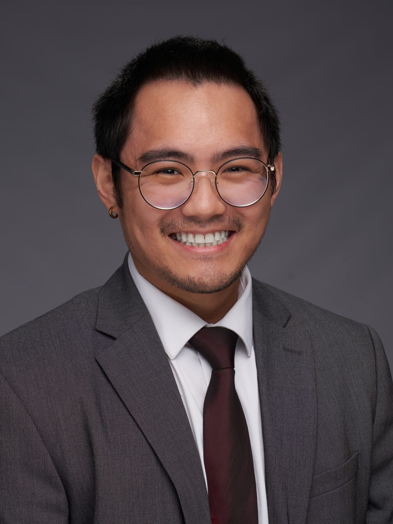 A person wearing glasses, a gray suit, white shirt, and dark red tie smiles at the camera against a plain gray background.