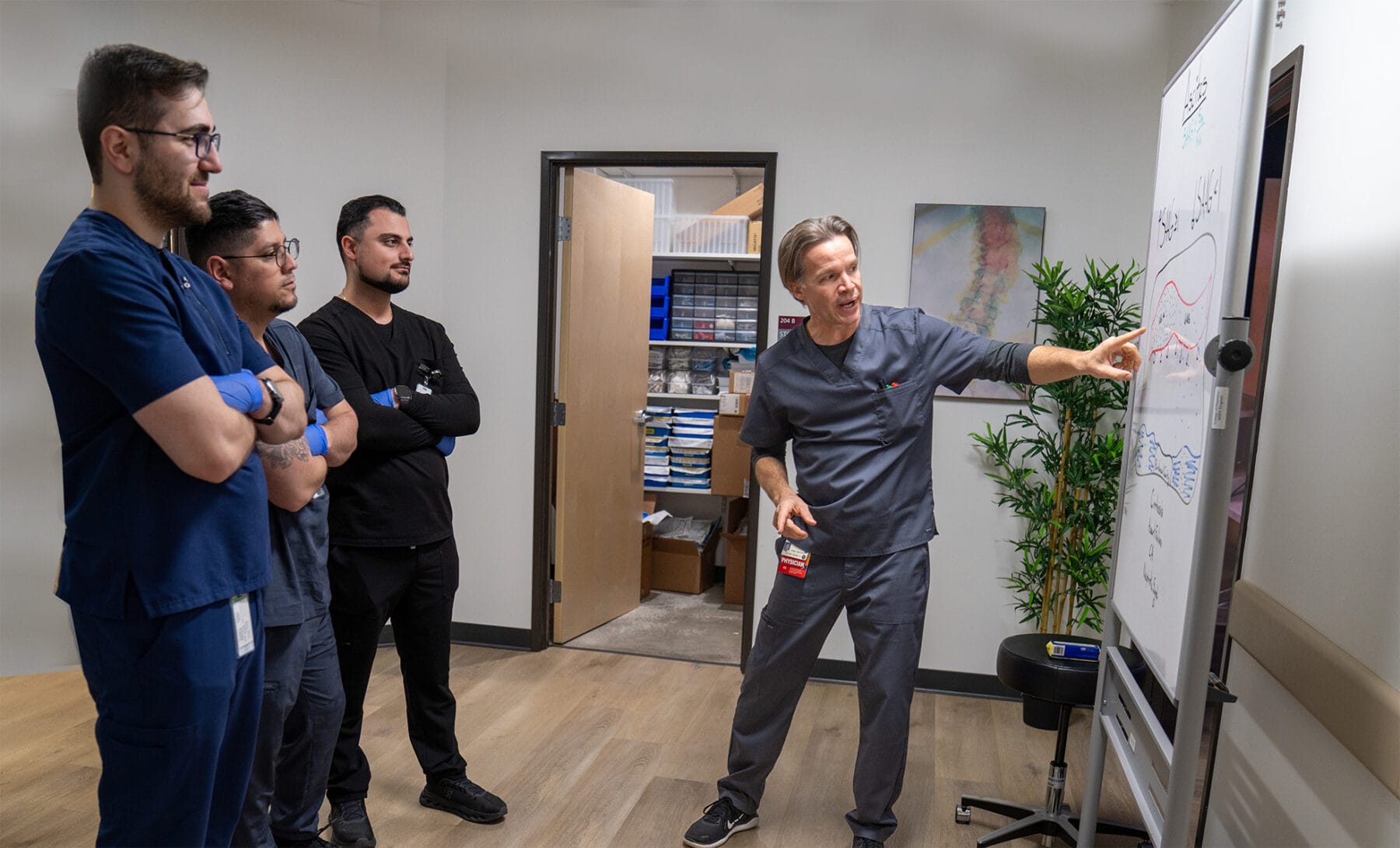 A man in medical scrubs points to a whiteboard with charts while three other people in scrubs watch attentively in a clinic office.
