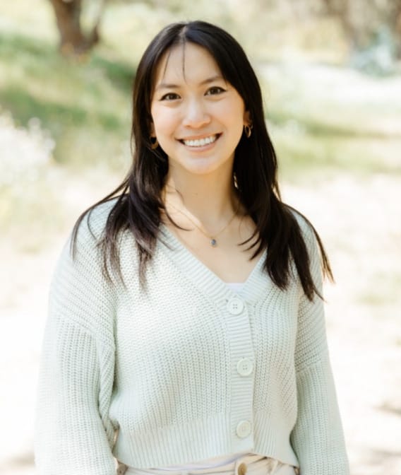 A woman with long dark hair wearing a light-colored knit cardigan stands outdoors, smiling at the camera with greenery in the background.