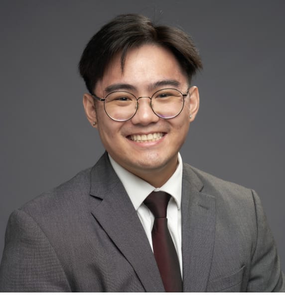 A young man with glasses, short dark hair, and earrings, wearing a gray suit, white shirt, and dark tie, smiling in front of a plain gray background.