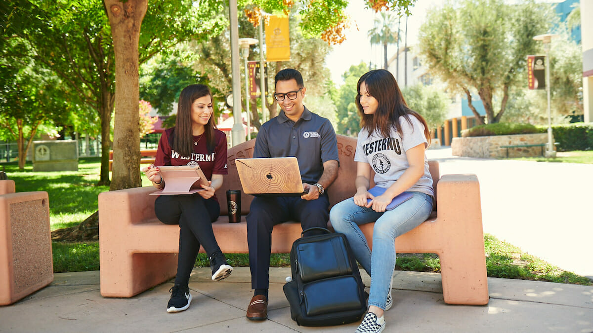 three students sitting on a bench with laptops.