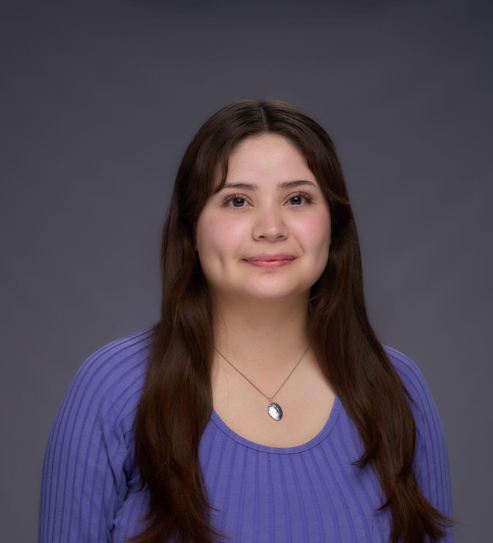 A woman with long brown hair wearing a purple top and a pendant necklace poses in front of a plain gray background.