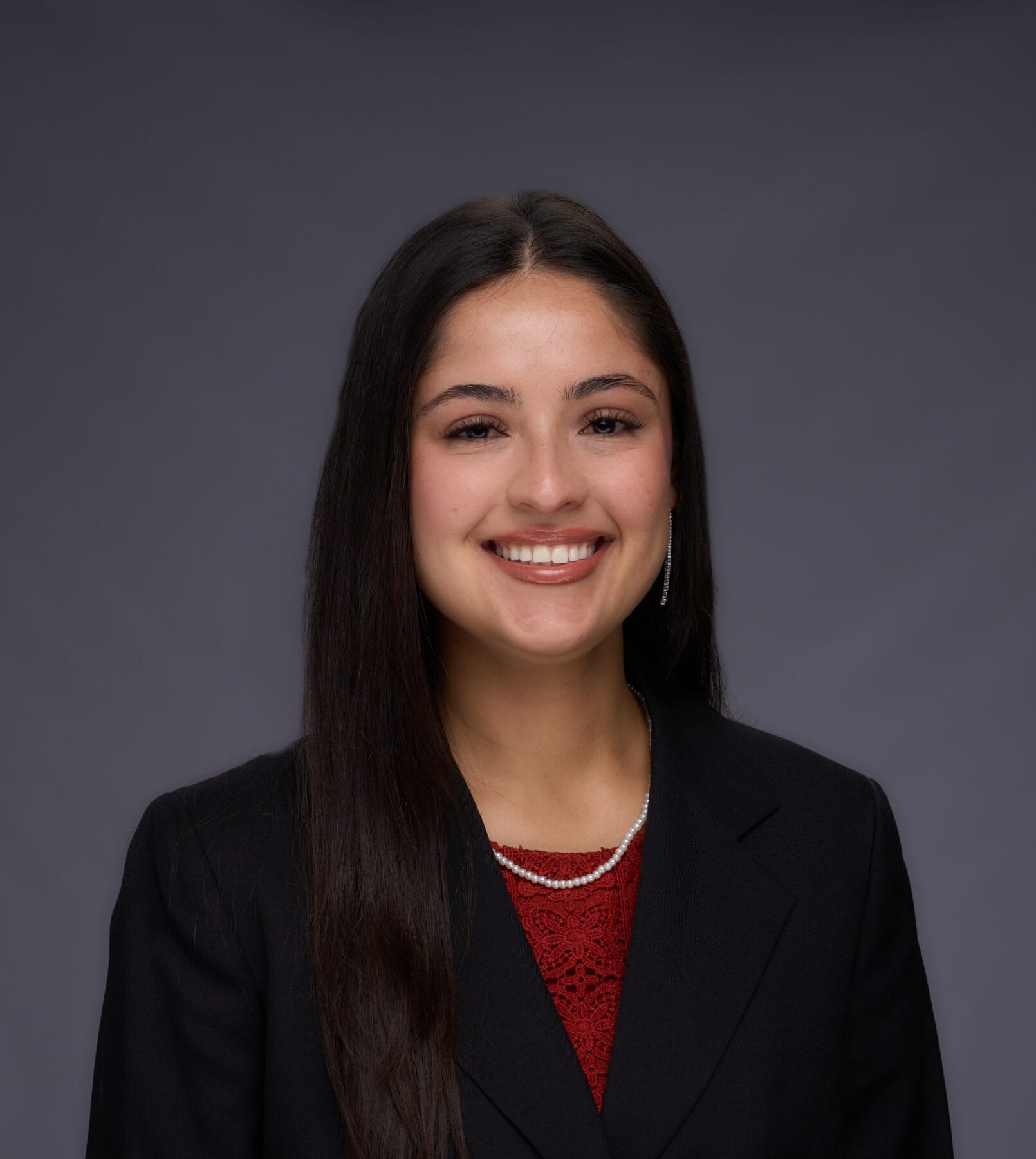 A woman with long dark hair, wearing a black blazer over a red top and a pearl necklace, smiles at the camera against a plain gray background.