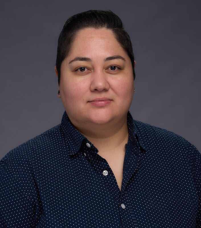 Person with short dark hair wearing a navy blue patterned shirt, facing forward against a plain gray background.