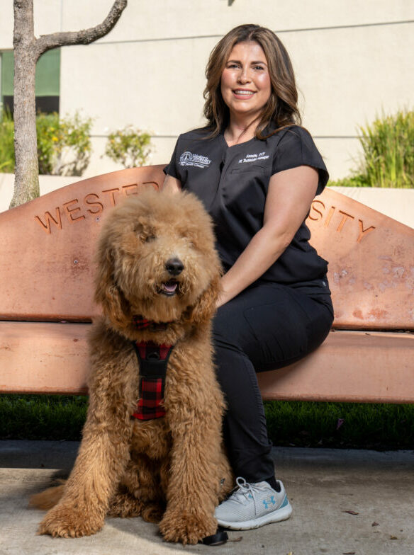 Veterinary Technician Manager Annette Chavarria-Marron holding a dog while sitting on a bench on the WesternU campus near a CVM building