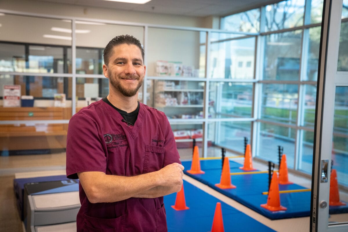 A man wearing maroon medical scrubs stands with folded arms in a room with agility equipment and orange cones.