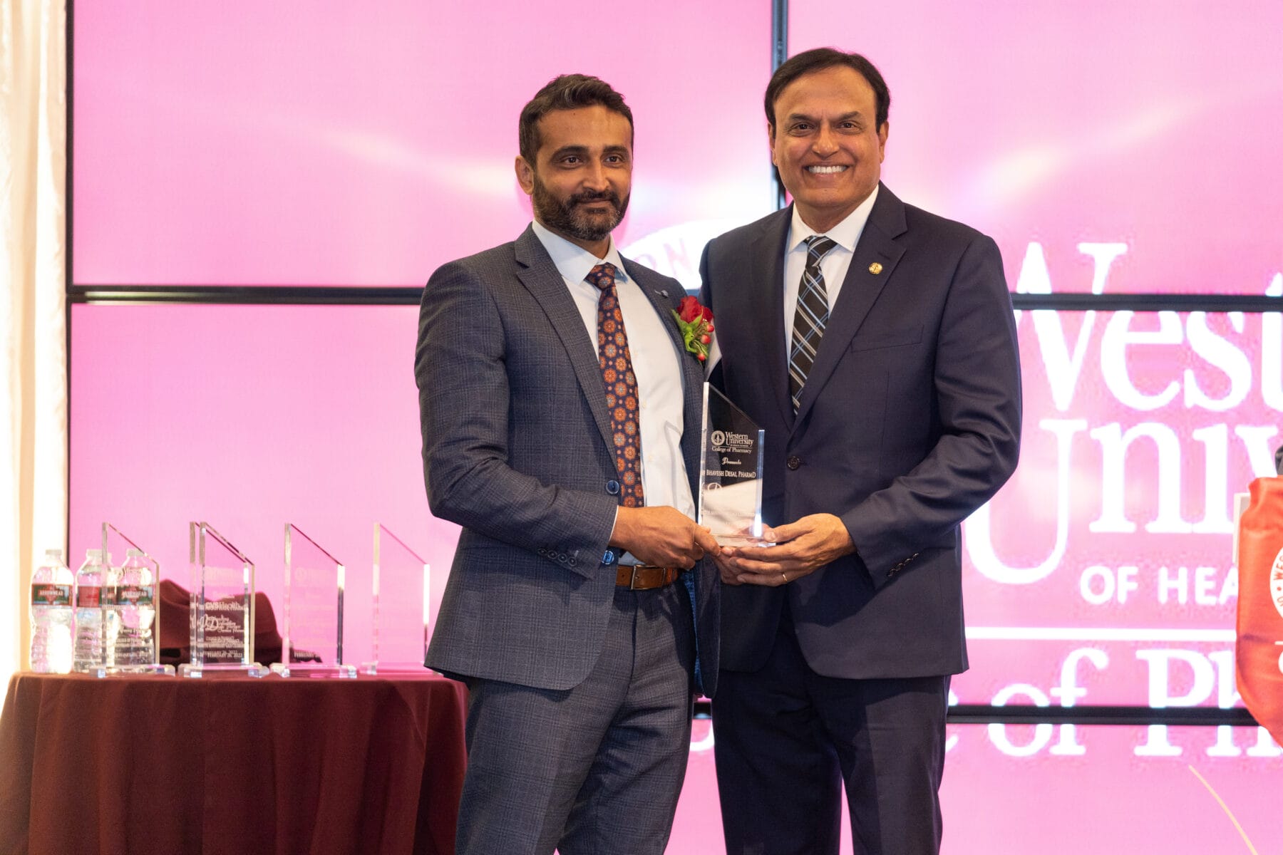 Two men in suits stand together holding an award on stage, with a table of trophies and water bottles beside them and a pink backdrop displaying text in the background.