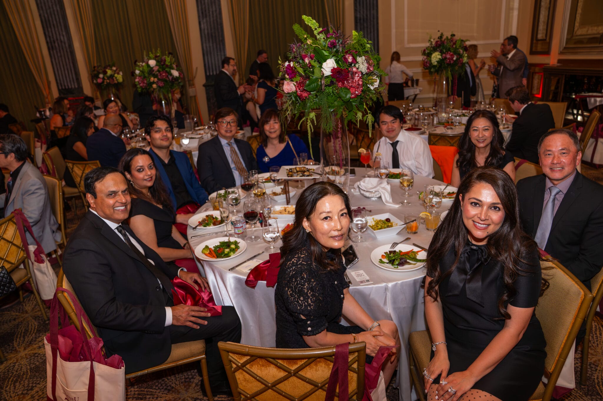 A group of people dressed formally sit around a round table with food and drinks at a banquet or gala event in an elegant venue.