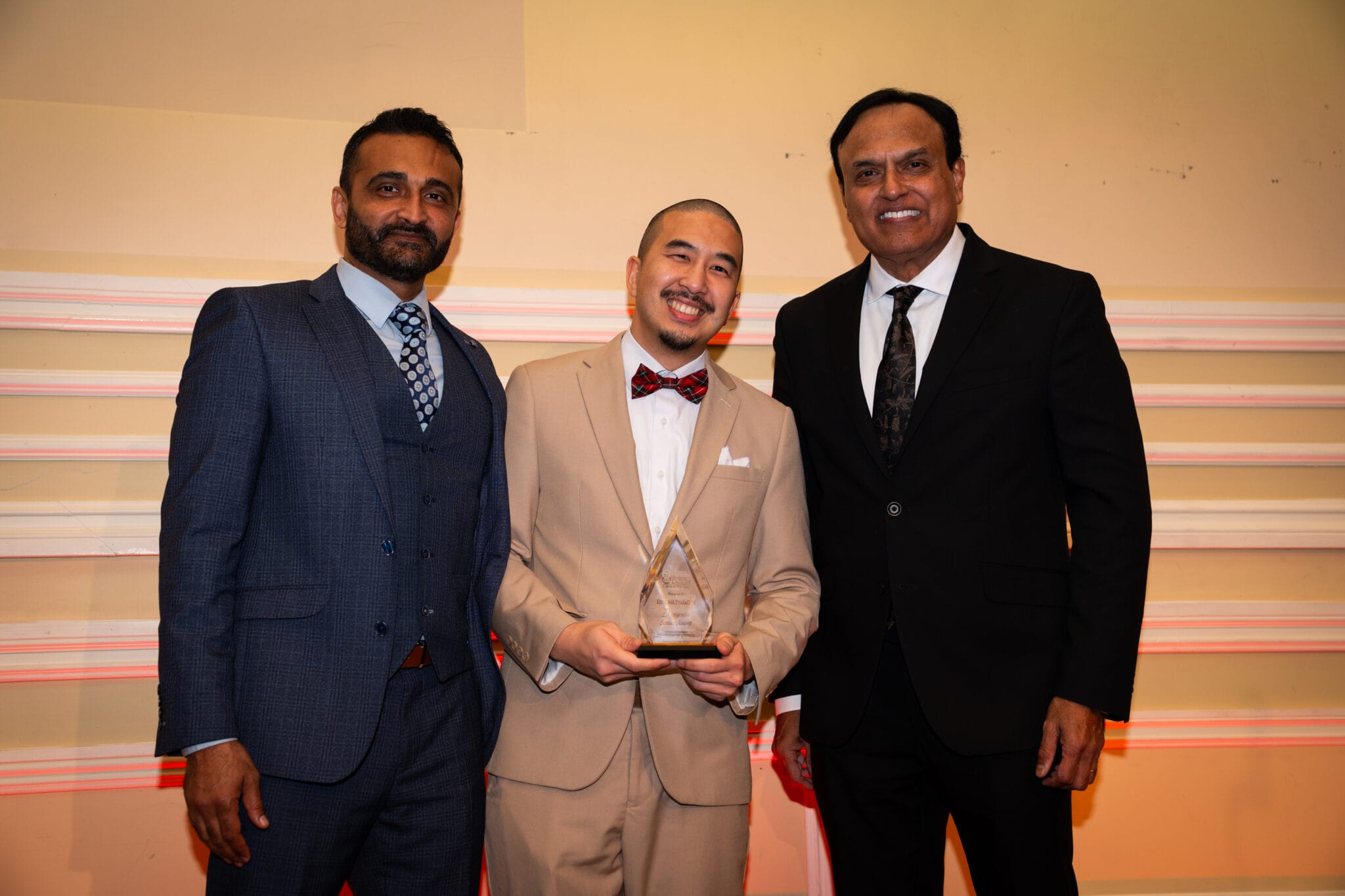 Three men in suits stand indoors; the man in the center holds a clear triangular award and smiles at the camera.